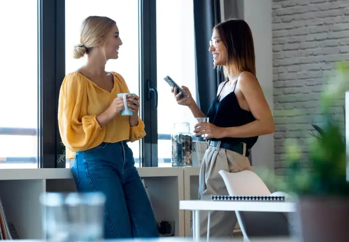 Dwie kobiety rozmawiają przy oknie, jedna z kubkiem, druga z telefonem. To przykład, jak wygląda **small talk co to** jest.