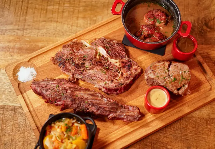 Un plateau en bois présente une entrecôte barbecue juteuse, des morceaux de viande braisée, des sauces et des légumes.