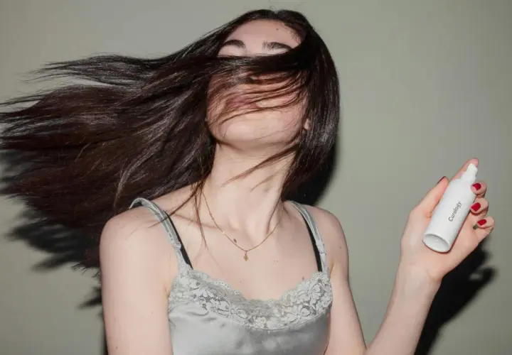 Mujer con cabello largo y oscuro moviéndose, sosteniendo un frasco de **aceite para el pelo**.