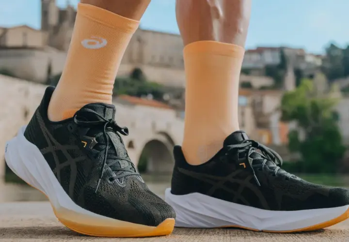Biegacz w pomarańczowych skarpetkach z logo firmy Asics i ciemnych butach sportowych na tle mostu i zabudowań.