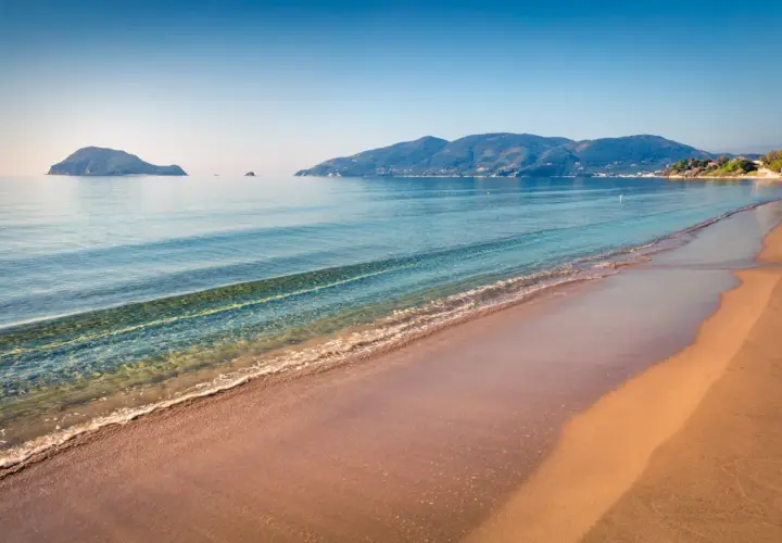 Spokojne morze obmywa piaszczystą plażę Kalamaki. W oddali widać zielone wzgórza i małe wysepki.