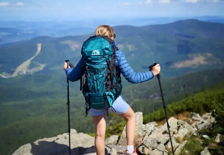 Kobieta z turkusowym plecakiem i kijkami trekkingowymi podziwia widok z góry. Idealny plecak turystyczny na wyprawę.