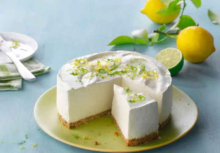 En krämig citron cheesecake med rivet citronskal som dekoration. Bredvid ligger färska citroner och en lime.