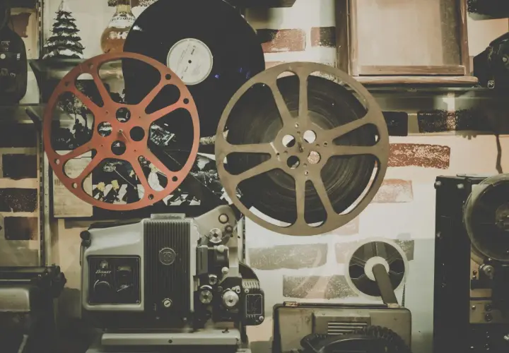Stary projektor filmowy, taśmy filmowe i płyta winylowa na tle ceglanej ściany.