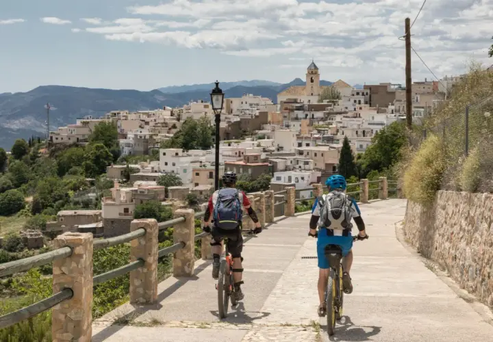 Dos ciclistas disfrutan de una ruta cicloturista ascendiendo hacia un pueblo blanco en la ladera de una montaña.