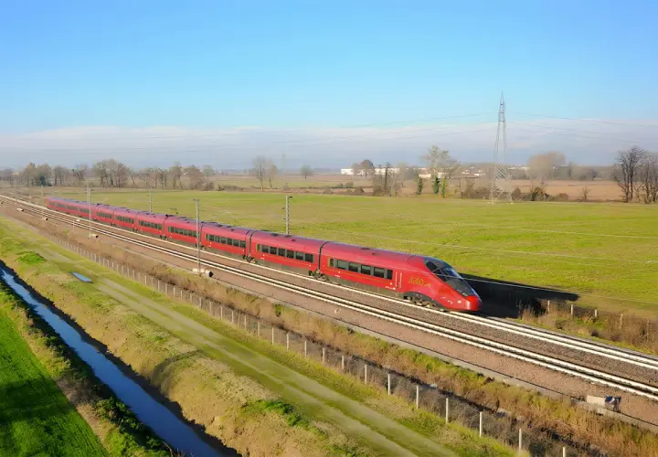 Le train rouge Italo file sur les voies, reliant Roma à Naples à travers la campagne verdoyante sous un ciel bleu.
