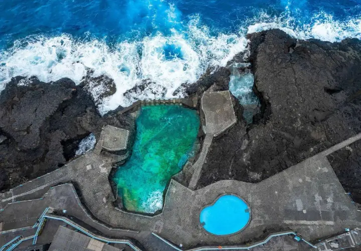 Piscinas naturales de agua turquesa y azul brillante, rodeadas de rocas volcánicas y el mar embravecido.