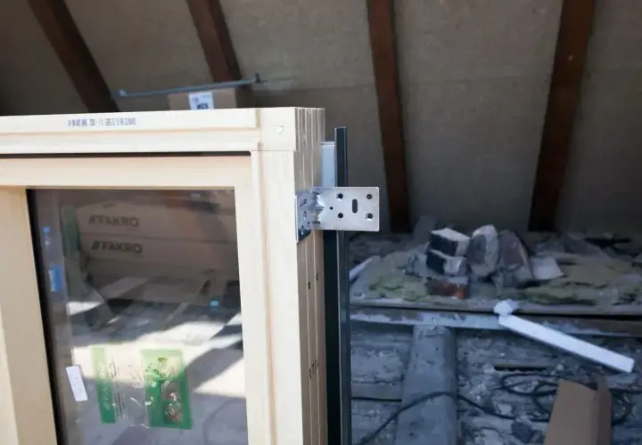 Drewniana rama okna z metalowym kątownikiem, gotowa do montażu. W tle widać materiały budowlane, co sugeruje obróbkę okien na poddaszu.