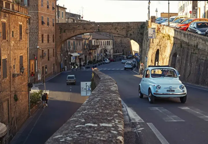 A light blue Fiat 500 drives through a Tuscan town. Do you need a car in Florence to explore charming streets like this?