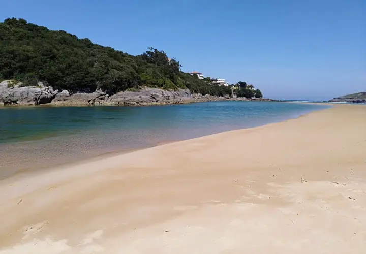 Amplia playa de la arena con aguas tranquilas y vegetación en la ladera. Un lugar ideal para los nudistas que buscan paz.