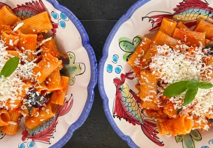 Deux assiettes de rigatoni alla norma, garnies de basilic frais et de fromage râpé, sur des plats décorés de poissons colorés.