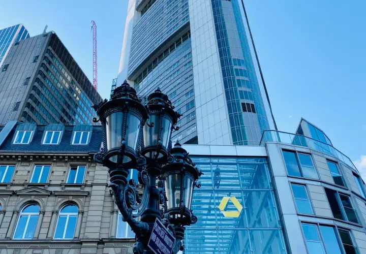Ein historischer Laternenpfahl vor dem modernen Commerzbank-Gebäude in Frankfurt. Die Fassade spiegelt den blauen Himmel wider.