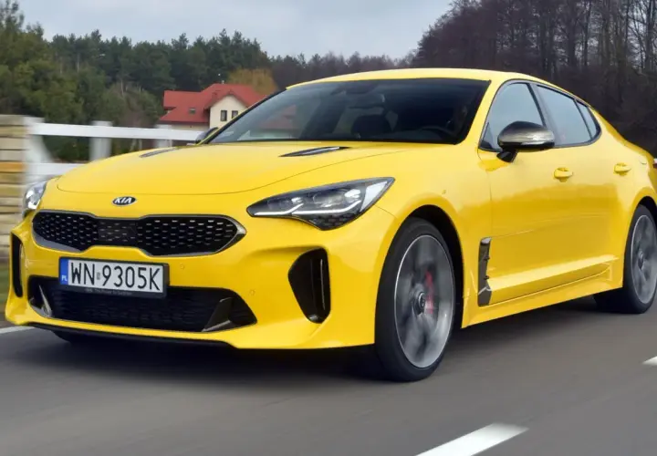 Żółty Kia Stinger GT pędzi po drodze, otoczony zielenią i domami.