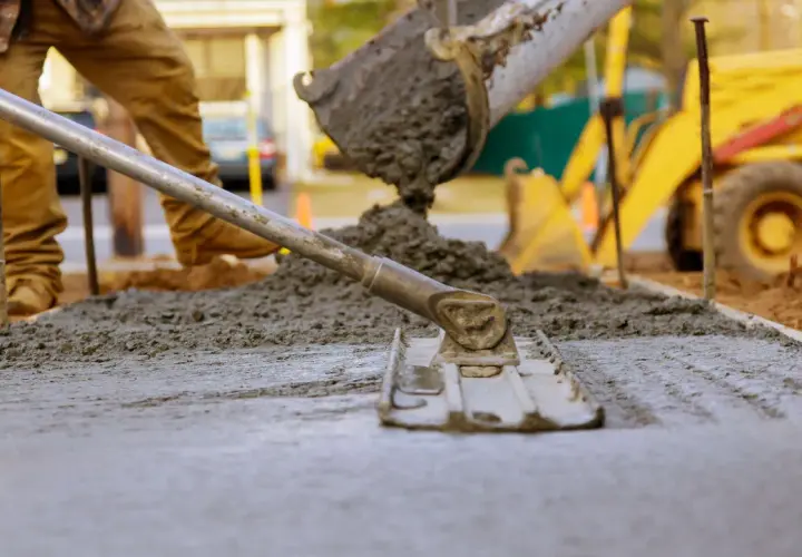 Pracownik wylewa beton na chudziaka, używając łaty do wyrównania powierzchni. W tle widać koparkę.