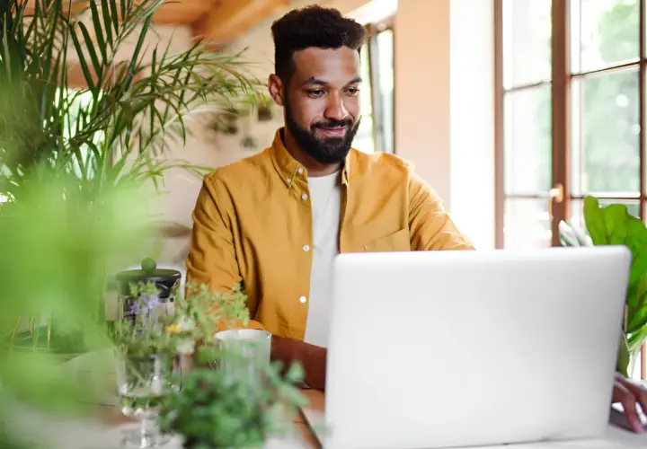 Uomo sorridente al laptop, circondato da piante. Sembra che stia imparando nuovi modi di dire per arricchire la sua comunicazione.