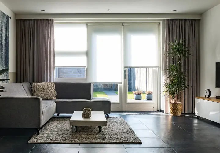 Grå soffa i vardagsrummet med draperade gardiner och stora fönster som släpper in ljus.
