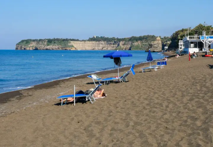 Plage de sable noir avec des chaises longues et des parasols bleus, face à une falaise rocheuse. Un doux air de Procida ou Capri.