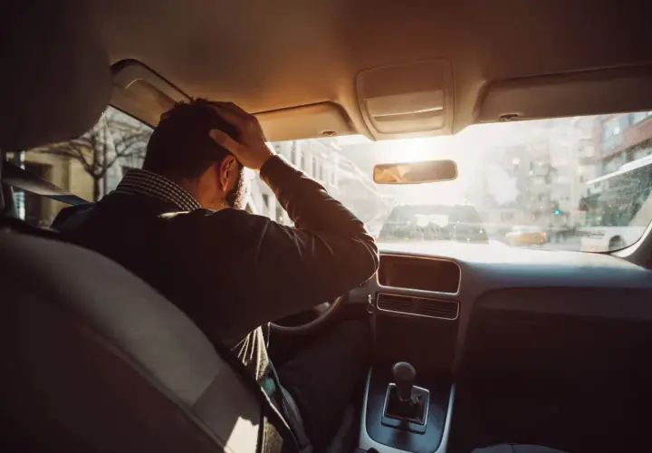 Hombre con mano en la cabeza dentro de un coche, buscando trucos para conducir sin nervios. El sol brilla a través del parabrisas.