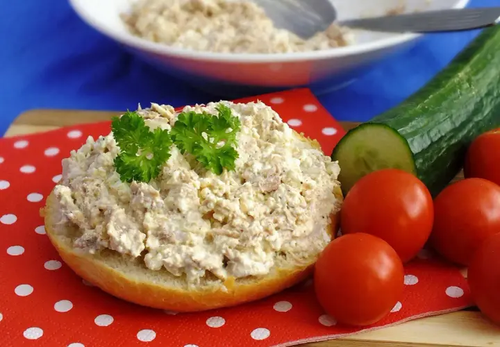 Pyszna pasta z tuńczyka i twarogu na bułce, ozdobiona natką pietruszki, obok pomidorki i ogórek.