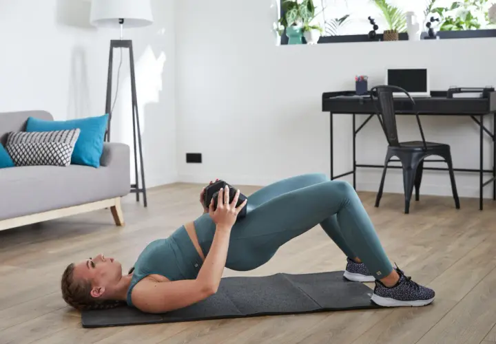 Kobieta wykonuje ćwiczenie glute bridge z obciążeniem na biodrach w domowym zaciszu.