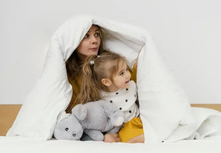Mama i dziecko bawią się pod białą kołdrą, która przypomina przytulne kołderki dla niemowląt. Obok leży szary pluszowy króliczek.