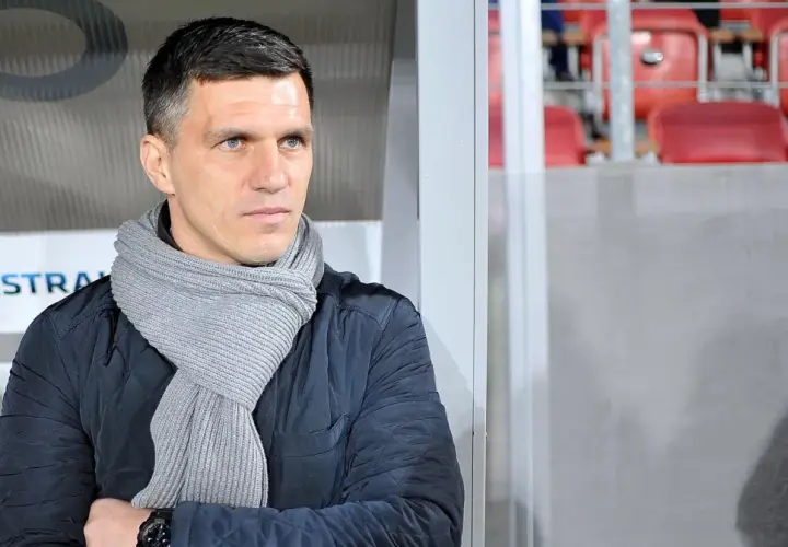 Robert Podoliński, trener w szaliku, spogląda w bok na tle stadionu z czerwonymi siedzeniami.