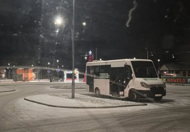 Biały bus na ośnieżonym przystanku nocą, ozdobionym świątecznymi lampkami.
