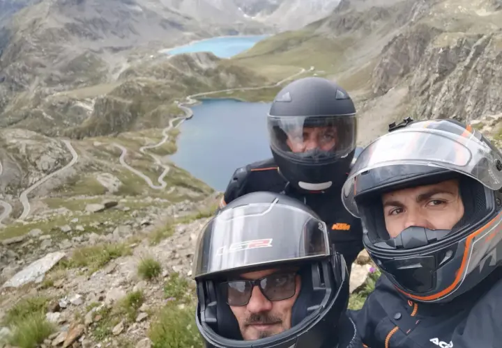 Tre motociclisti con caschi ammirano il panorama mozzafiato del Col del Nivolet, con i suoi laghi alpini e le strade tortuose.