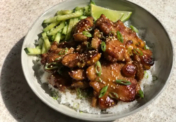 En skål med ris, gurka och saftig grillad kyckling med sesamfrön och salladslök. En limeklyfta ligger bredvid.