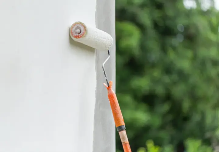 Fassade streichen ohne Reinigung: Ein Malerroller mit orangefarbenem Griff überzieht eine weiße Wand mit Farbe. Im Hintergrund verschwimmen grüne Bäume.