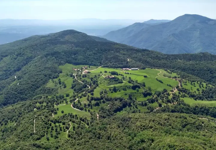 Vista aérea del **santuari del far**, rodeado de verdes colinas y bosques frondosos. Caminos serpentean entre prados y edificios dispersos.
