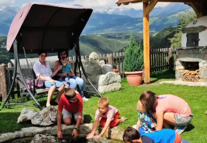 Famiglia rilassata in un giardino con vista sulle montagne, tipico delle fattorie altoatesine. Bambini giocano vicino a un laghetto, mentre adulti riposano su un'altalena.