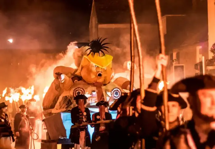 Un pupazzo gigante con un cappello giallo, circondato da fiamme e persone in costume, celebra un giorno festivo in inglese.