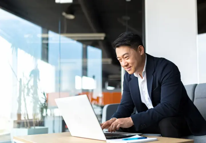 An Asian man in a suit smiles while working on his laptop, perhaps studying for his bachelors in organizational leadership.