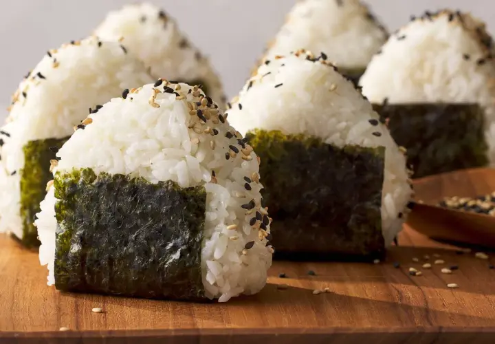 Several triangle sushi onigiri, sprinkled with sesame seeds and wrapped in nori, sit on a wooden board.