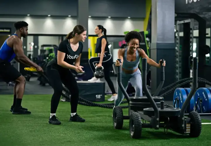 Intensywny trening interwałowy na siłowni. Kobieta pcha sanie, mężczyzna ćwiczy z linami, a inni wykonują ćwiczenia z kettlebell.