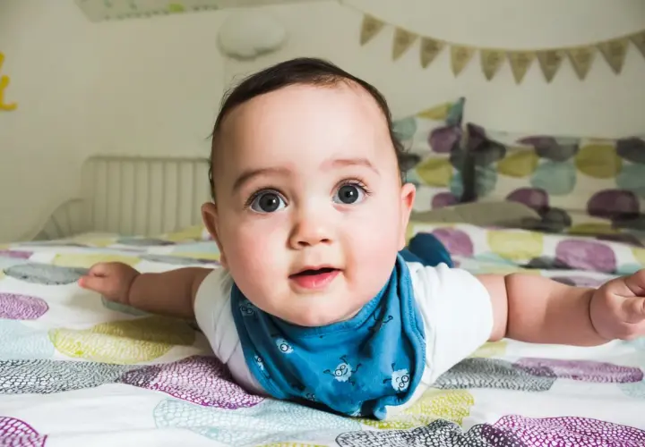 Un adorabile neonato di 4 mesi, sdraiato a pancia in giù sul letto, con gli occhi spalancati e un bavaglino blu.