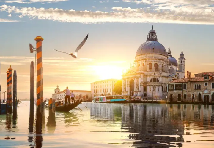 Góndola navega por canal veneciano al atardecer, con la Basílica de Santa Maria della Salute y otras **ciudades de Europa** reflejadas en el agua.