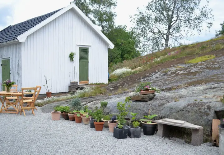 Vitt hus med gröna detaljer och ett bord med stolar. En grusgång leder fram till huset, omgiven av sten och växter.