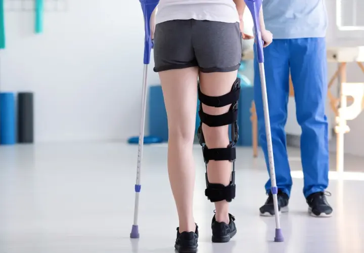 Rehabilitacja po złamaniu nogi: pacjentka w ortezie i z kulami ćwiczy pod okiem fizjoterapeuty.