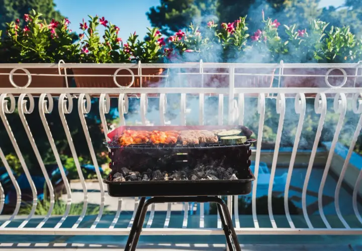 Grill z mięsem i warzywami na balkonie, w tle kwiaty i błękitne niebo.