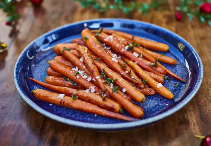 Zanahorias al horno, tiernas y doradas, espolvoreadas con sal marina y hierbas frescas, servidas en un plato azul sobre una mesa de madera.
