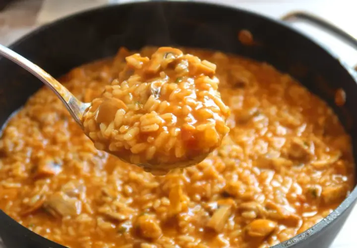Cuchara sirviendo arroz caldoso con pollo y champiñones, humeante y apetitoso, de una sartén.