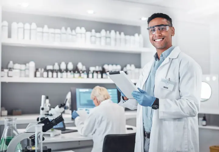 Uśmiechnięty technik farmacji w okularach ochronnych trzyma tablet w laboratorium, gdzie na półkach stoją liczne butelki, a w tle pracuje inna osoba.