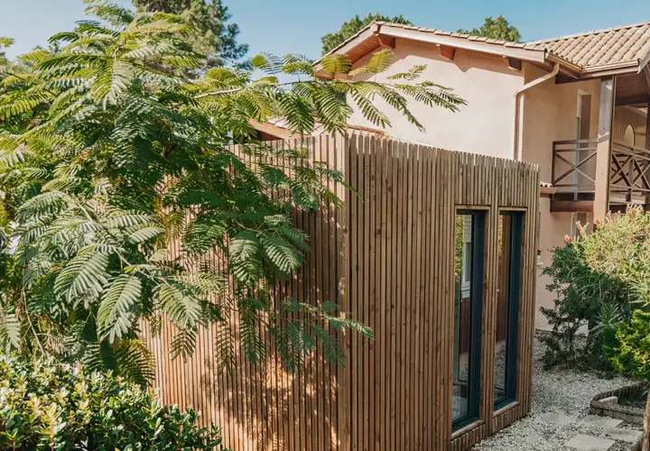 Extension de maison en bois moderne, bardage vertical, fenêtres sombres. Un espace de vie supplémentaire sans permis de construire, intégré dans un jardin verdoyant.