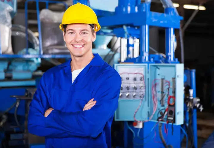 Uśmiechnięty pracownik w żółtym kasku i niebieskim kombinezonie, stojący przed panelem elektrycznym. To może być pracownik hurtowni elektrotechnicznej Comel Rumia.