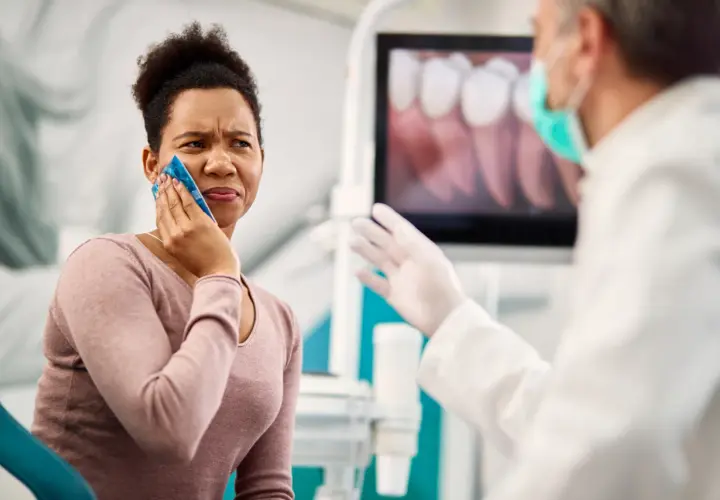 Mujer con compresa fría en la mejilla, sufriendo un alveolitis con dolor insoportable, mientras el dentista le explica algo.