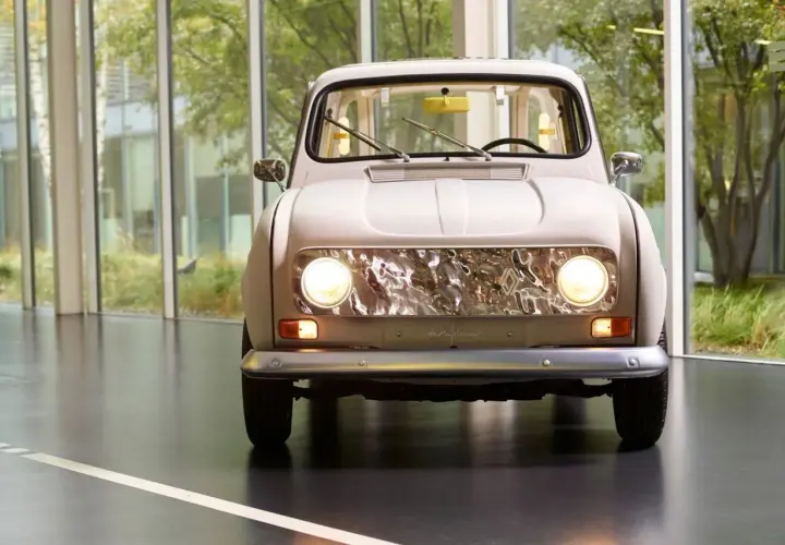 En ljusbeige Renault 16 står framför stora fönster med grönska utanför. Bilens front med sina runda strålkastare och kromade grill är i fokus.