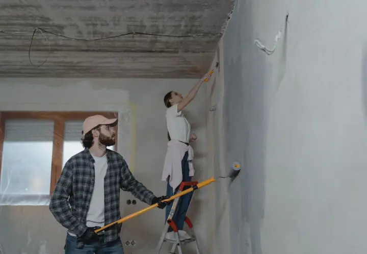 Para robi poprawki na pomalowanej ścianie. Mężczyzna z wałkiem w ręku, kobieta na drabinie maluje sufit.