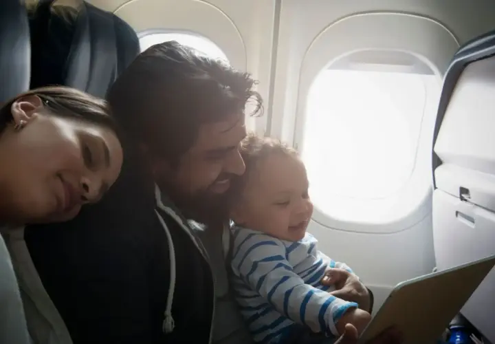 Familj flyger med bebis. Pappa håller ett leende barn som tittar på en surfplatta, mamman sover bredvid.